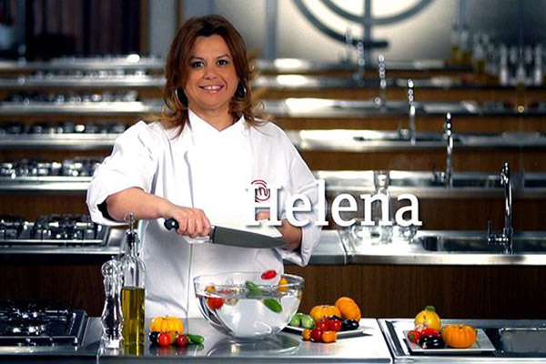 Helena Manosso Helena Manosso sorrindo atrás de bancada do MasterChef enquanto despeja legumes cortados de tábua para tigela de vidro.