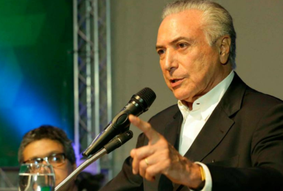 Michel Temer aparece fazendo um discurso, com o microfone preto à frente e o dedo indicador estendido