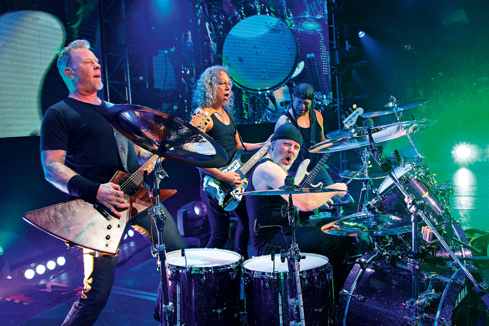 Imagem mostra três homens com seus instrumentos em um palco iluminado. Um guitarrista, um baixista e um baterista