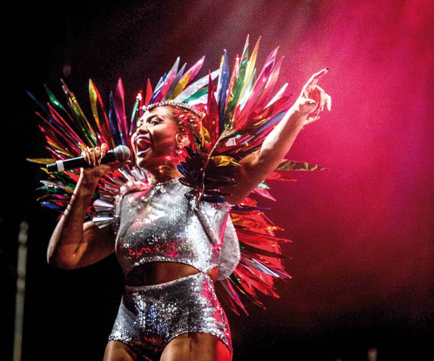 Foto de Gaby Amarantos em show, segurando microfone, com a boca aberta e usando roupa com fitas coloridas