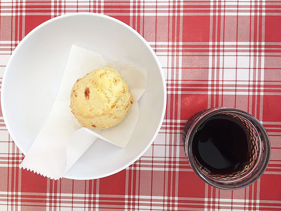 Taça de vidro com sorvete sobre base redonda de madeira, com pedaço de goiabada, pedaço de queijo e pequena faca ao lado, sobre mesa de madeira branca