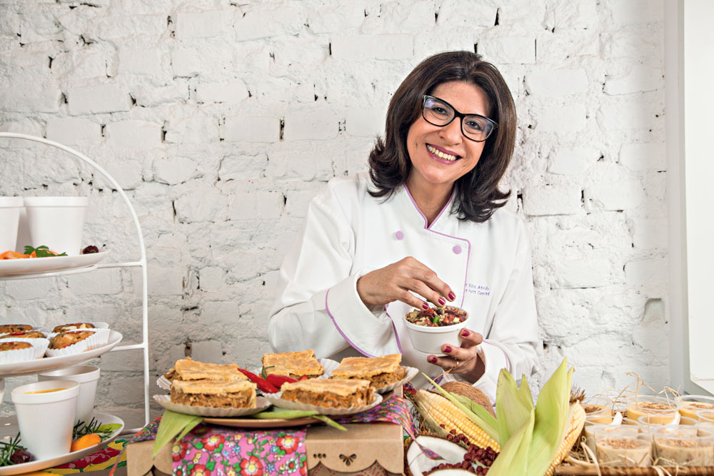 A chef Rita Atrib posa de óculos e roupa branca em frente a mesa com quitutes.