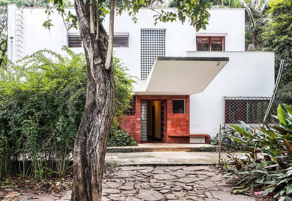 Foto mostra fachada da Casa Modernista, com árvore à esquerda e portas duplas de madeira ao fundo, com parede branca com cobogós acima