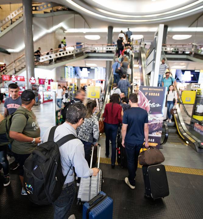 Foto mostra passageiros com malas andando em direção a escada rolante