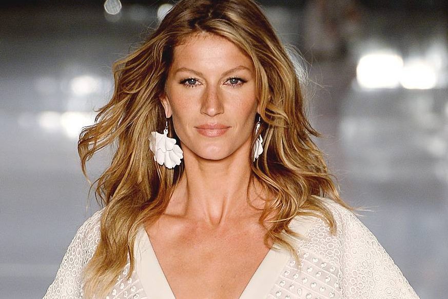 Modelo Gisele Bündchen na passarela com cabelos em movimento, brincos brancos e roupa branca.