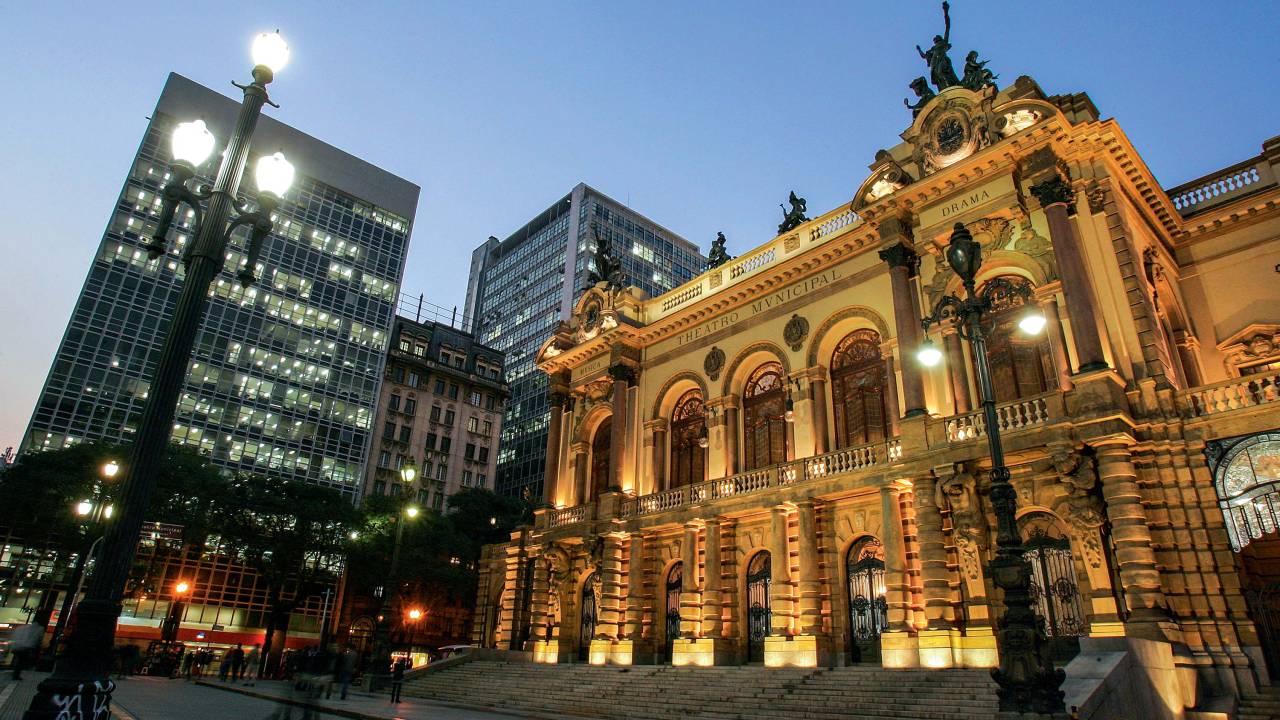 A imagem mostra a faixada do Teatro Municipal, iluminada, em um fim de tarde com o céu parcialmente escuro.