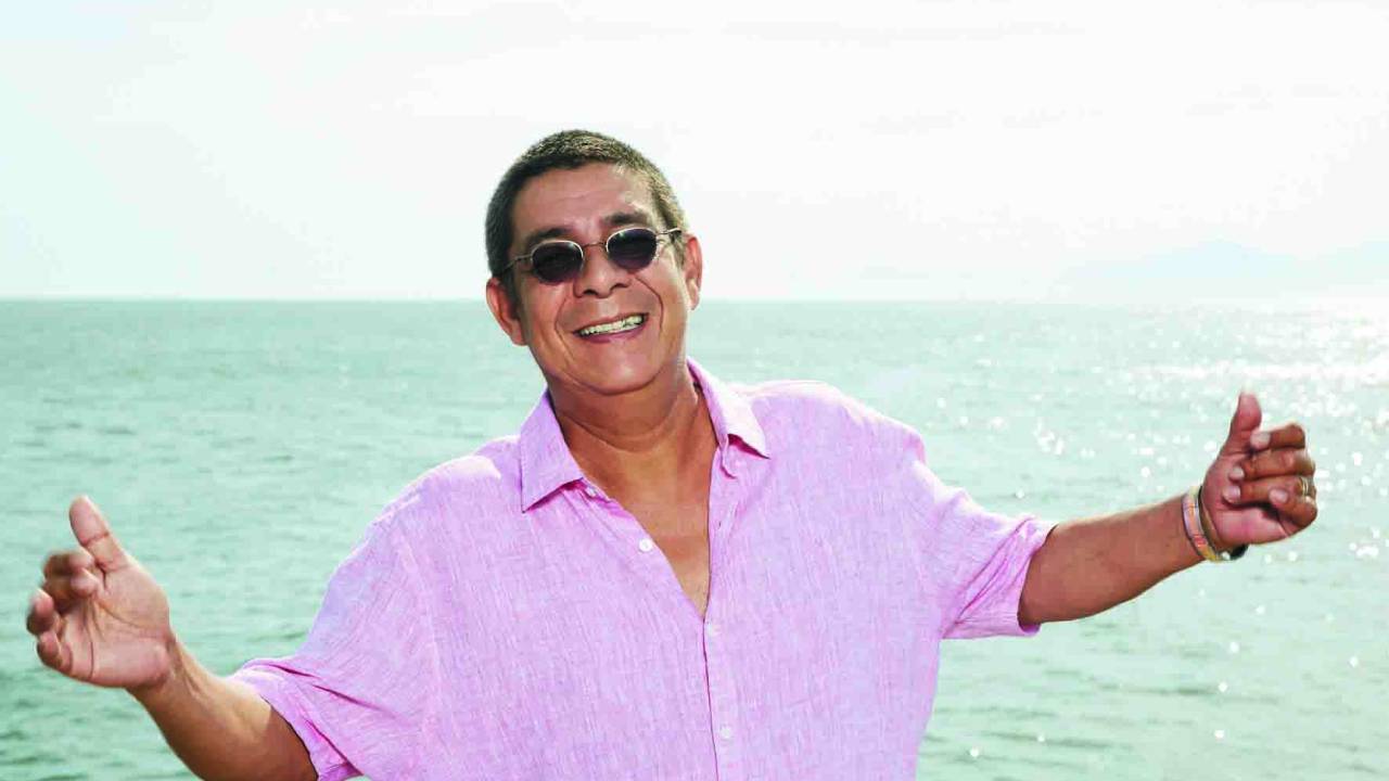 Imagem mostra Zeca Pagodinho sorrindo, usando óculos, na frente do mar