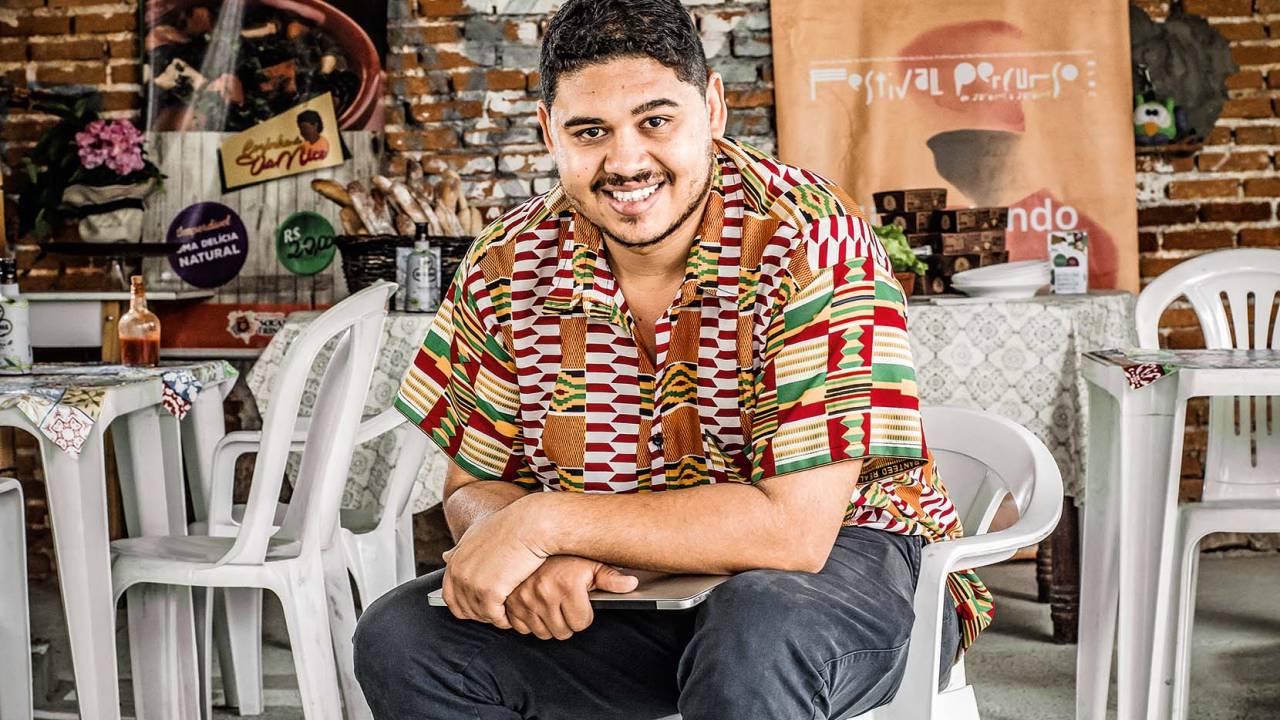 Homem sorrindo ao centro vestido com camisa de manga curta colorida e calça jeans escura, sentado em cadeira de plástico.