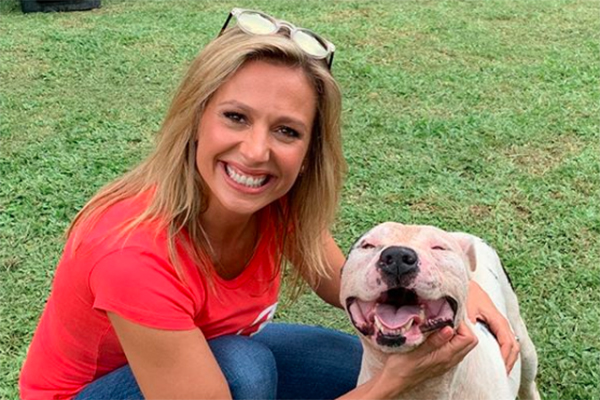 Imagem mostra Luísa Mell ao lado de cachorro, sorrindo; os dois estão agachados em um gramado, Luísa usa óculos, camiseta e calça jeans