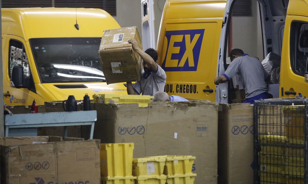 Um homem aparece segurando uma caixa de papelão sobre o ombro. Ao fundo, carros da Sedez