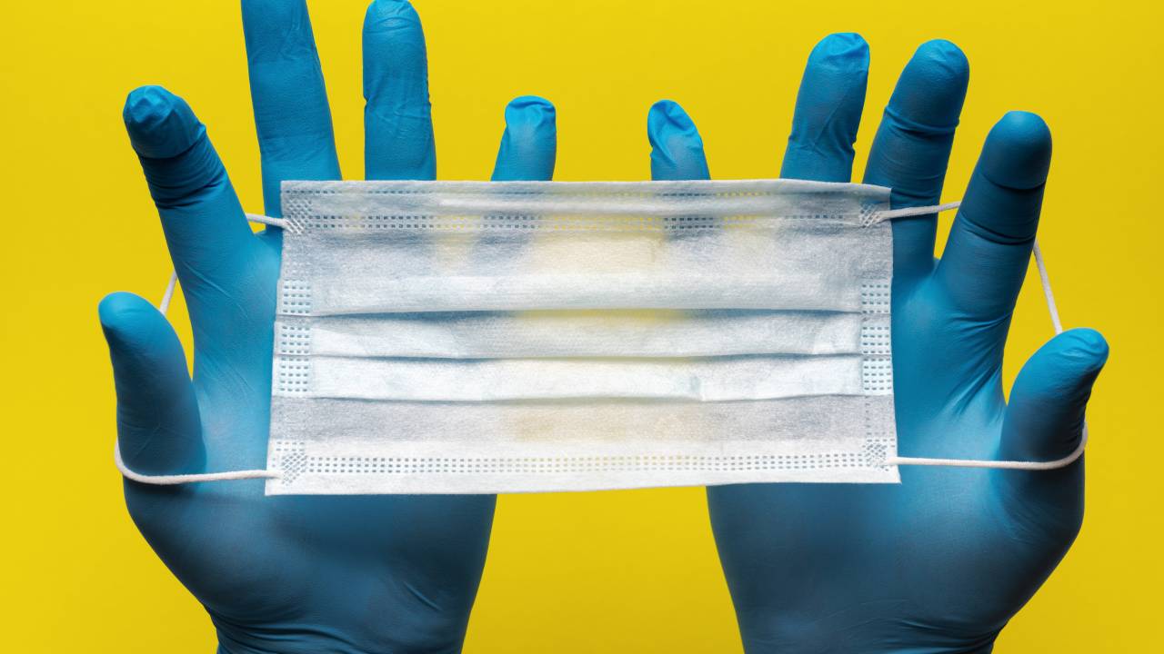 Imagem mostra pessoa com par de luvas cirúrgicas segurando máscara