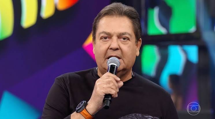 Imagem mostra Faustão apresentando o Domingão; ele usa relógio e segura microfone, enquanto olha para a câmera