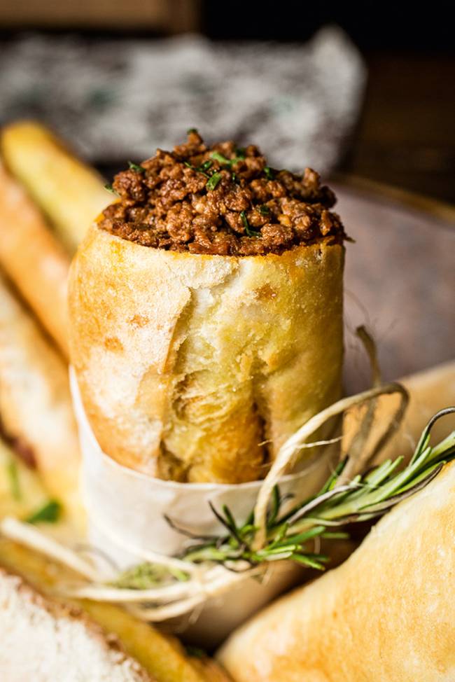 Pão francês em pé preenchido de carne moída ao lado de ramos de alecrim.