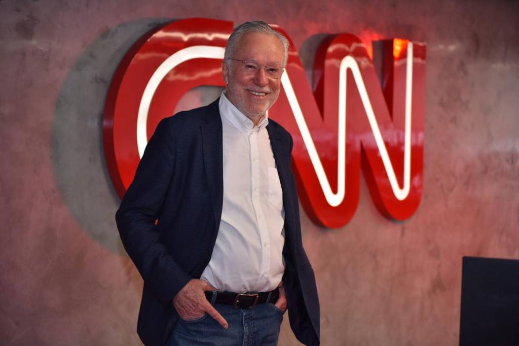 A imagem apresenta Garcia, em frente ao logo da CNN, com uma roupa social e sorrindo para a câmera