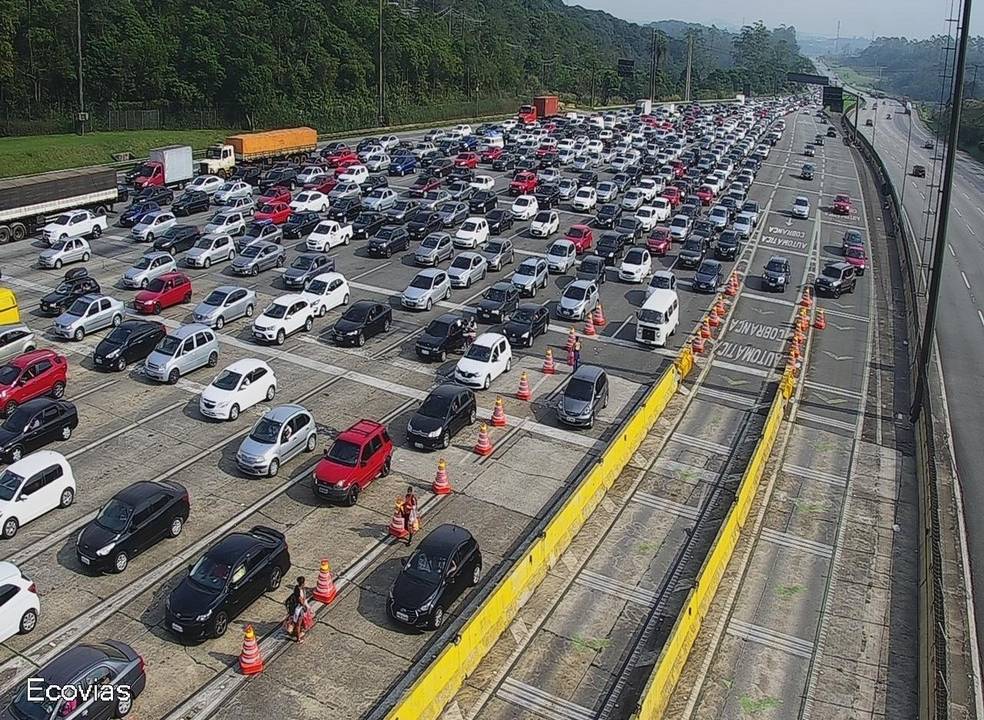 Imagem de pedágio exibe fileiras de carros em direção ao litoral.