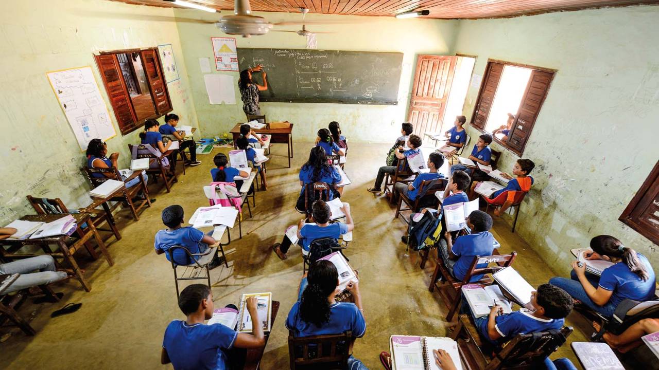 Vista de cima, foto exibe sala de aula com alunos vestidos de azul e quadro negro ao fundo.