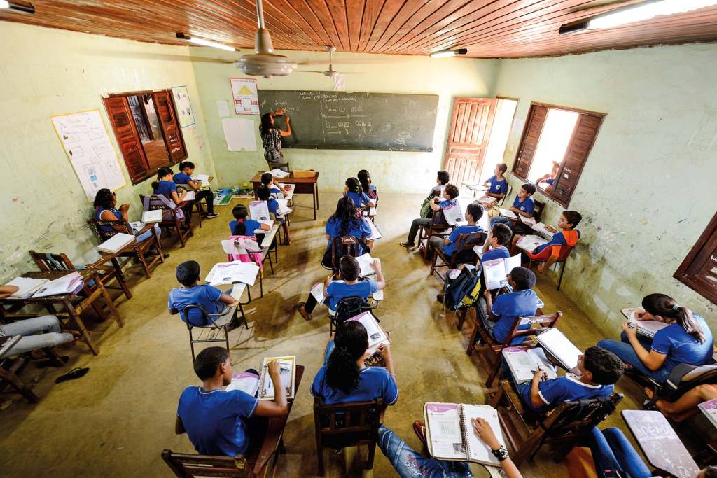 Vista de cima, foto exibe sala de aula com alunos vestidos de azul e quadro negro ao fundo.