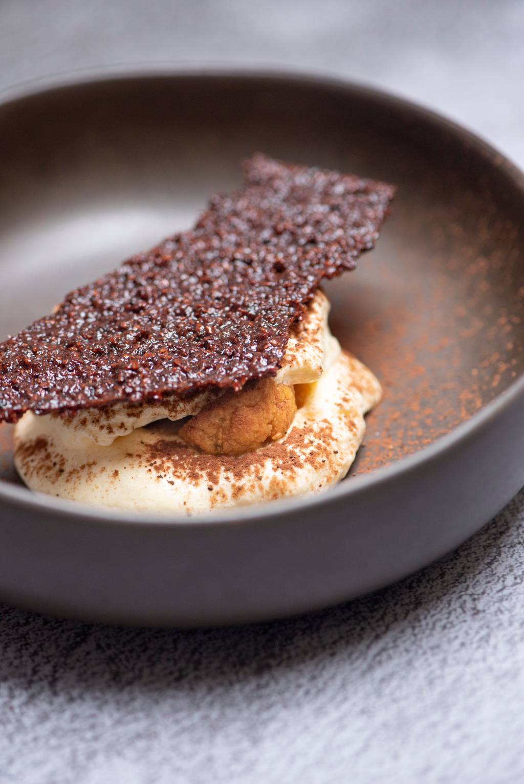 Tiramisu do Fame coberto de telha de nibs de cacau em prato fundo de cerâmica marrom.