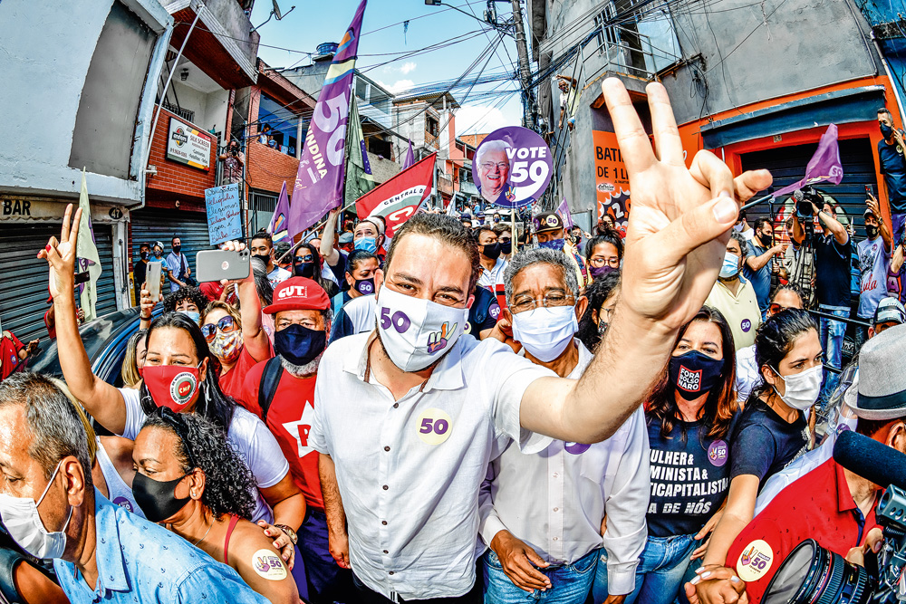 Guilherme Boulos em aglomeração em Heliópolis. Na imagem ele está em meio a muitas outras pessoas, com um braço levantado e fazendo um sinal com os dedos indicadores e médio para cima