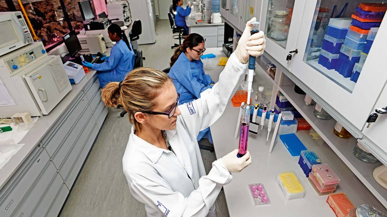 Imagem de uma mulher em um laboratório, equipada com um jaleco branco e óculos de proteção