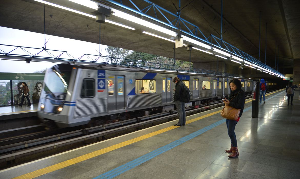 Um trem do metrô parada em estação. Há duas pessoas esperando na plataforma.