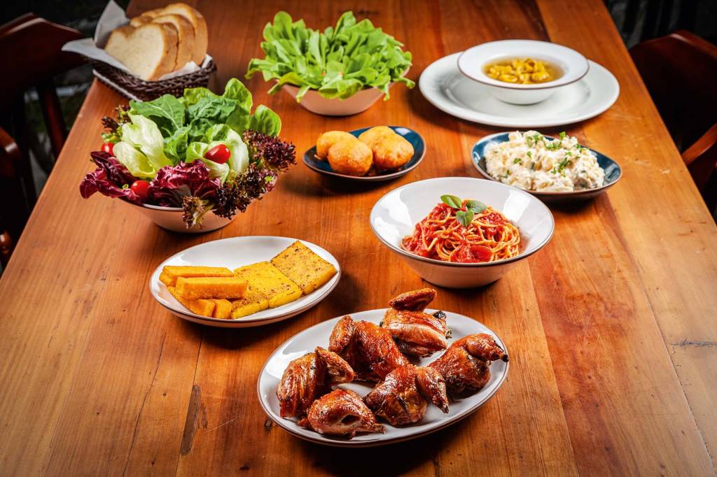 Comer & Beber 2020/2021 – Bom e Barato – Restaurantes – Galeto Di Paolo Rodízio do Di Paolo sobre mesa de madeira. Com galetinhos em travessa na frente com polenta frita atrás, prato fundo com macarrão, saladas, bolinhos e sopa.