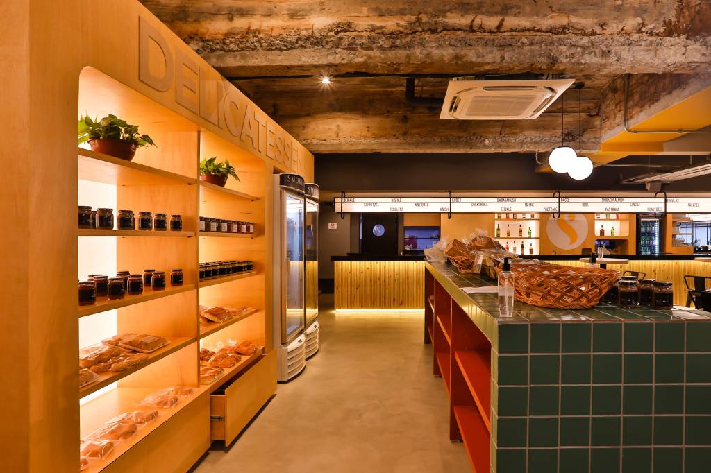Smokedeli (Luciano Braz) (3) Prateleiras exibem produtos kosher iluminados e alinhados com balcão verde e vermelho à direita, em ambiente de empório da lanchonete.