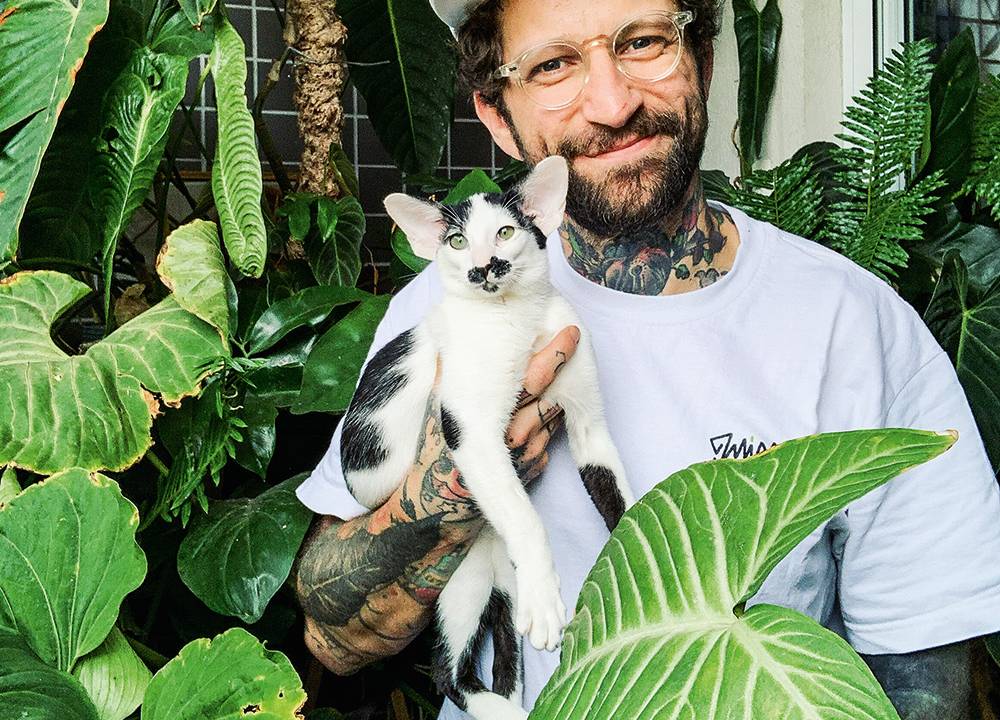 Taniel posa de boné e camisa branca, com barba e óculos de aros redondos, e o gato de estimação Roberto em um dos braços. Ao seu redor, plantas de folhas verdes enchem o cenário.