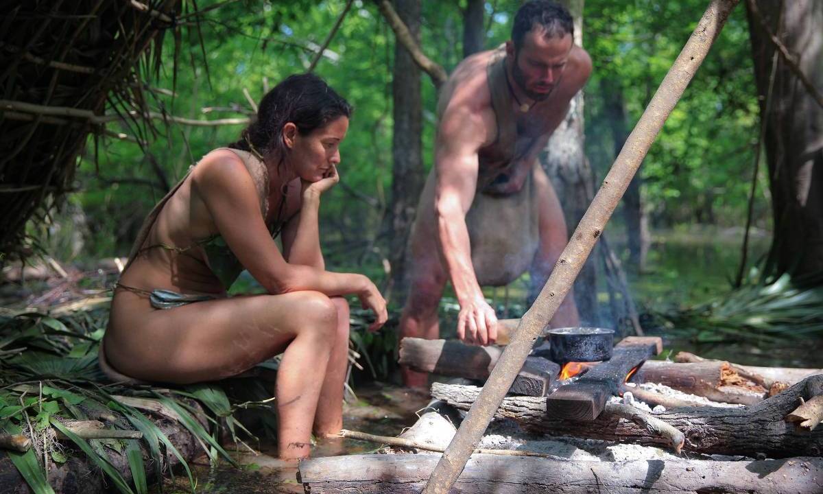 Uma mulher e um homem, vestidos com peças de roupas curtas que parecem primitivas, acendem uma fogueira no meio da floresta