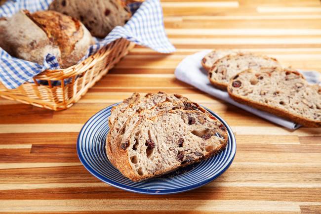 Fatias de pão sobre prato azul ao centro, em cima de mesa de madeira. No canto superior esquerdo, cesta de palha com pães e à direita, guardanapo de pano dobrado com três fatias de pão.