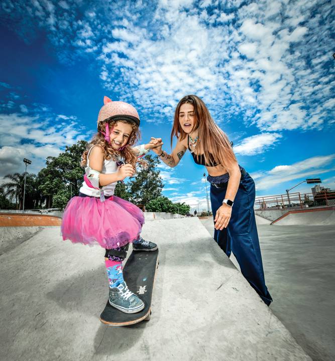 Uma menina equipada com proteção andando de skate de mãos dadas com uma mulher
