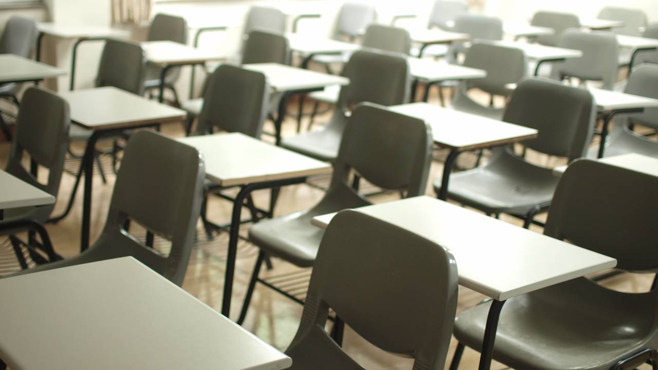 Imagem de carteiras escolares enfileiradas e vazias dentro de sala de aula