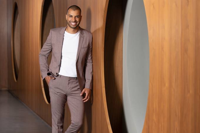 Wellington Melo, novo diretor do Unique, posa de terno cor bordô, tênis brancos e camiseta branca, encostado em parede cor de madeira com grandes círculos vazados.