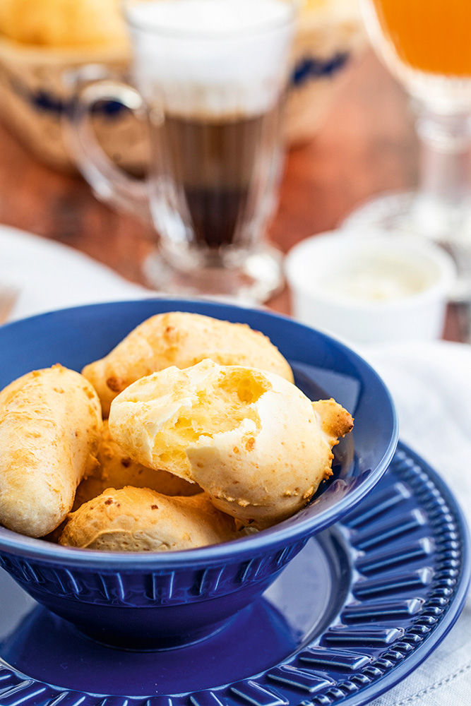 Taça de vidro com sorvete sobre base redonda de madeira, com pedaço de goiabada, pedaço de queijo e pequena faca ao lado, sobre mesa de madeira branca