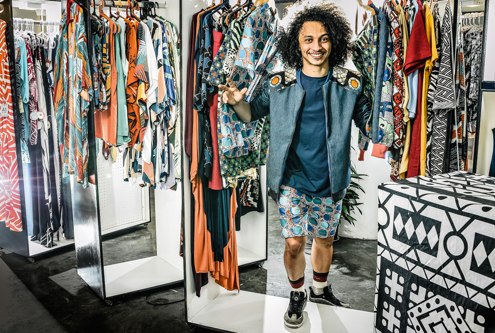 Sandro Freitas sorrindo para a foto entre as roupas de sua marca