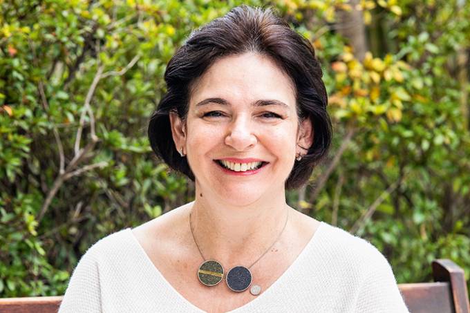 Eliz Taddei, sorrindo para a câmera, sentada em um banco de madeira.