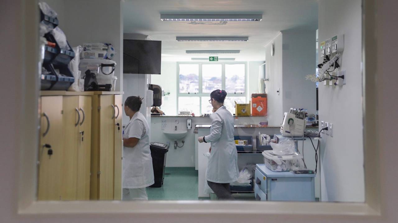Na imagem é possível ver uma sala de hospital em que há duas mulheres vestindo equipamentos médicos, como jaleco