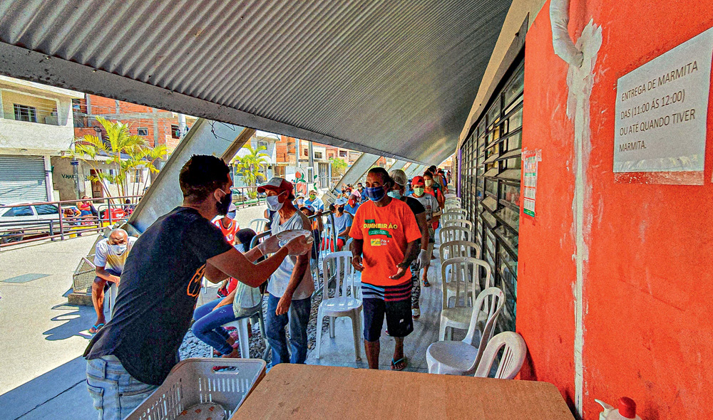 Homem, de máscara, entregando marmitas em uma fila ao ar livre