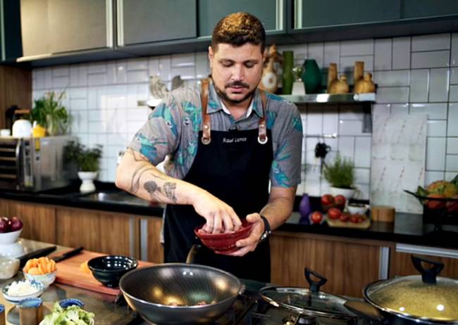 Raul Lemos Homem com avental culinário jogando ingredientes de uma cumbuca para uma frigideira funda numa cozinha