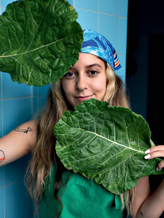Luísa Motta mulher segurando folhas de couve nas mãos próximas ao rosto, uma no queixo e outra na testa