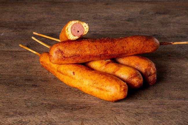 Três corn dogs juntos sobre superfície de madeira com mais um espeto por cima e outra cortado ao meio, mostrando a salsicha no recheio, sobre todos.