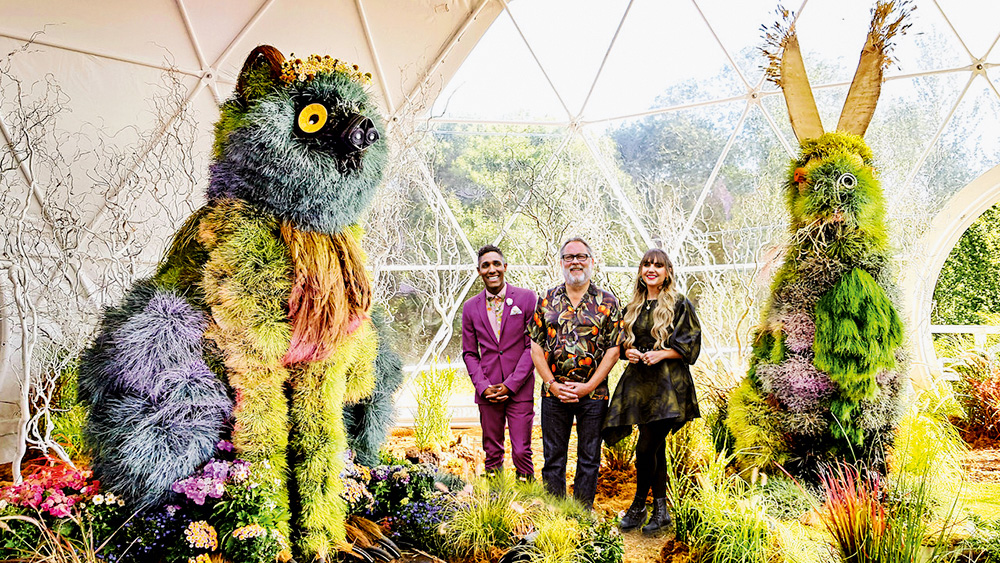 Dois homens e uma mulher bem arrumados posam em um jardim super ornamentado, com flores e plantas gigantes