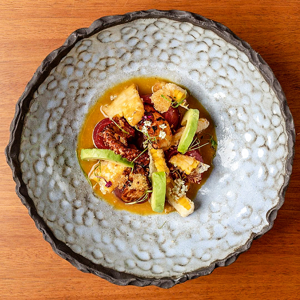 Imakay – Tiradito tierra y mar Com fundo de madeira, prato aparece em primeiro plano com os ingredientes: polvo, atum, abacate e tempurá de milho.