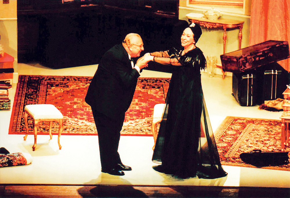 Alta Sociedade – 003 (1).JPG Na imagem, Sergio e Fernanda estão de pé em cima de um palco, vestidos com roupas chiques. Ele está inclinado, segurando as duas mãos da atriz, com a cabeça próximo delas, enquanto ela olha para cima sorrindo.