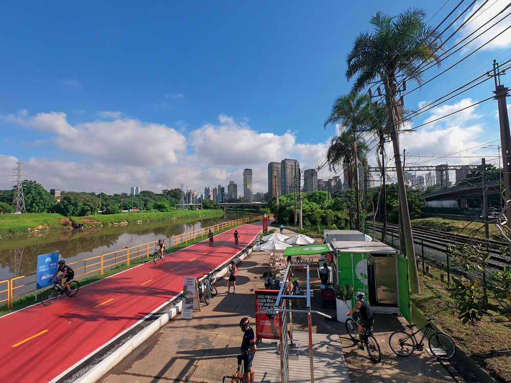 Imagem vista de cima da ciclo faixa do Rio Pinheiros. Há ciclistas de capacete utilizando a pista em um dia com céu azul com apenas nuvens ao fundo