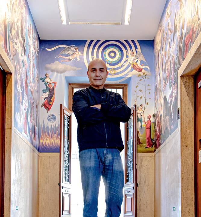 Artista plástico posa de bracos cruzados no corredor principal do Colégio Dante Alighieri. Veste calças jeans, blusa preta de manga longa. Ao fundo, trecho do mural aparece com cenas da obra A Divina Comédia, em tons azuis e dourados.