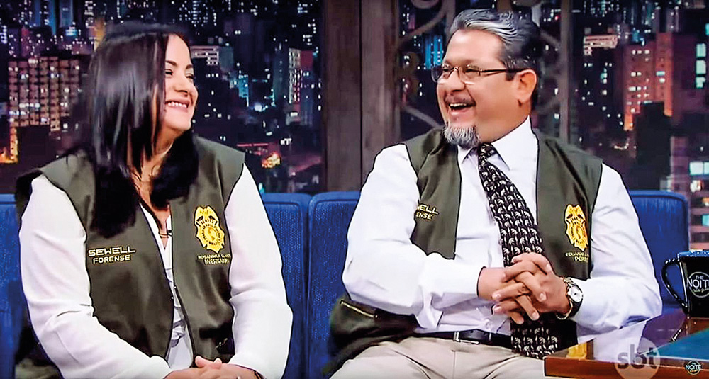 Entrevista casal yt Eduardo e Rosangela sentados em um sofá azul, vestidos com uniforme de criminalistas. Eles estão sorrindo. No fundo há um cenário de um programa de talkshow.