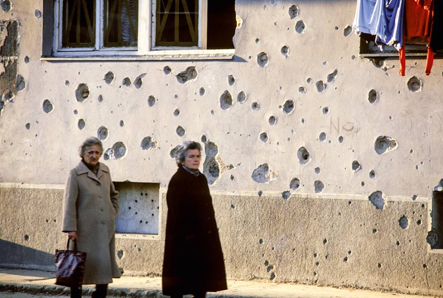 Marcas de bombardeios nas paredes e carro destruído abandonado na rua: cidade-ruína e ecos da violência