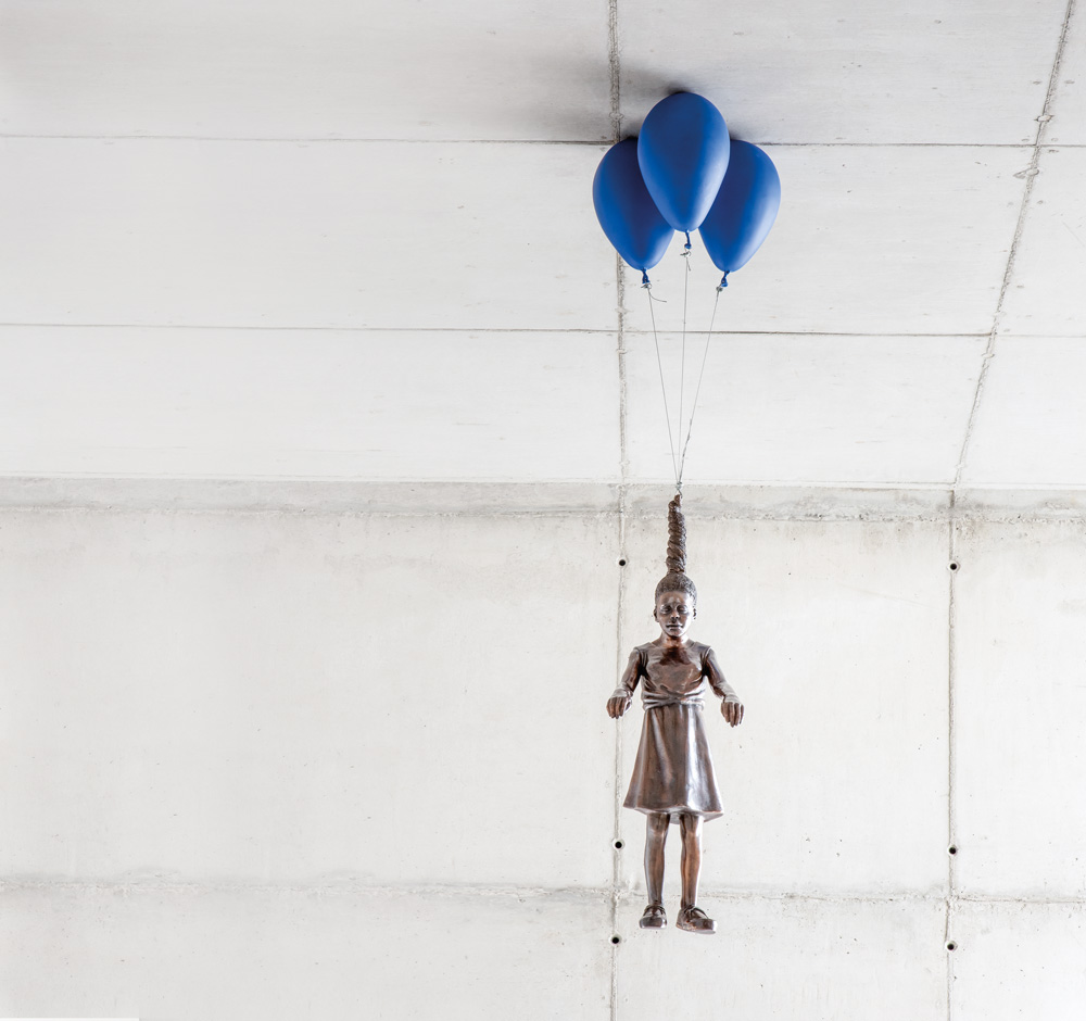 Escultura de Flávio Cerqueira em bronze com uma menina de pendurada no teto da Galeria Leme por três bexigas