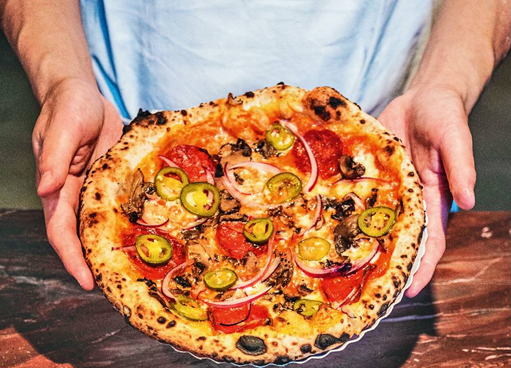 Uma pessoa segurando uma pizza individual sobre um balcão de madeira